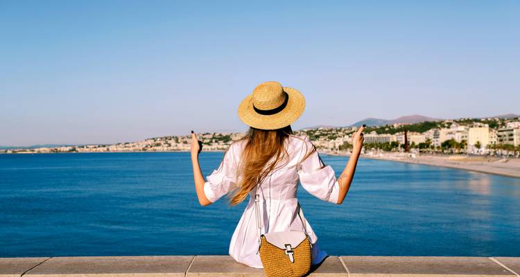 Mujer en vestido de verano y sombrero de paja disfrutando de una vista panorámica de la ciudad costera desde un paseo marítimo.