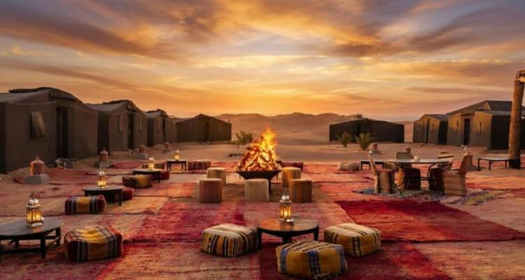 Patio de campamento en el desierto al atardecer con una fogata ardiente, mesas bajas, alfombras con patrones y faroles contra cielos resplandecientes