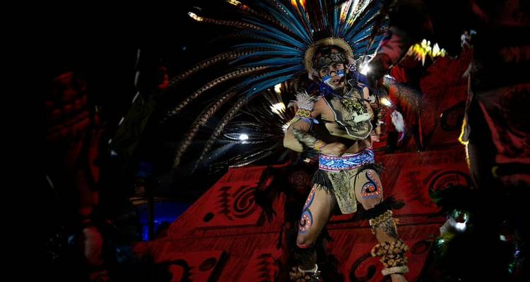 Energieke danser in Azteekse stijl met gevederd kostuum verlicht op een donker podium.