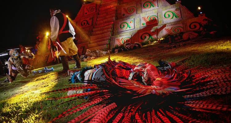 Nachtelijke theaterscène met uitvoerders rond een piramidedecor, levendig rood en geel licht.