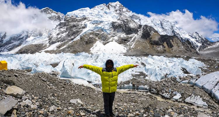 Trekker in een felgele jas strekt armen wijd uit te midden van ijs en rotsen op Everest Base Camp met torenhoge besneeuwde pieken boven het hoofd.