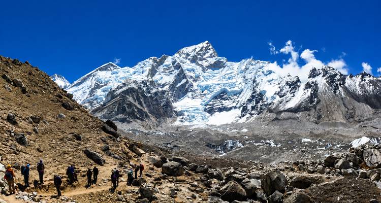 Rij trekkers die over een rotsachtig pad wandelen richting massieve met ijs bedekte Himalaya-toppen onder een diepblauwe hemel.