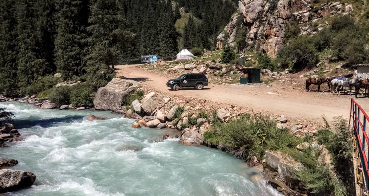 Río turquesa caudaloso junto a un sendero de tierra que conduce a un campamento de yurtas en un valle de coníferas.