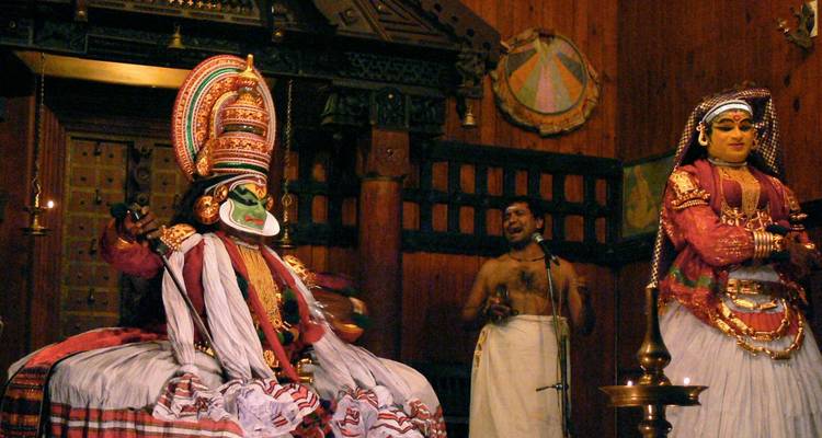 Traditionelle Kathakali-Tänzer in aufwendigen Kostümen treten auf einer Holzbühne auf, während ein Sänger sie begleitet.