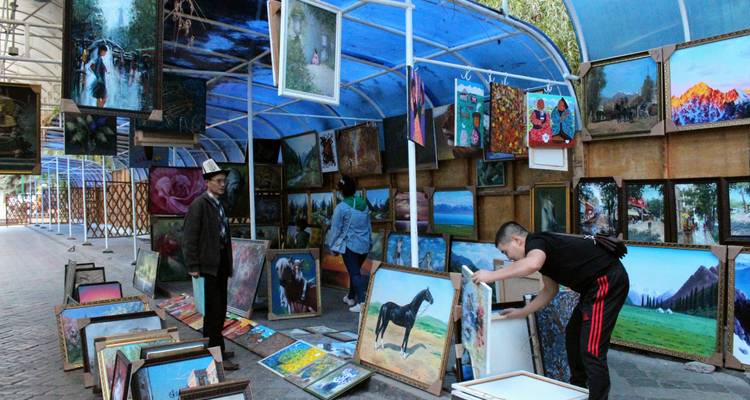 Mercado de arte al aire libre con numerosas pinturas enmarcadas exhibidas bajo toldos azules mientras vendedores y compradores las observan.