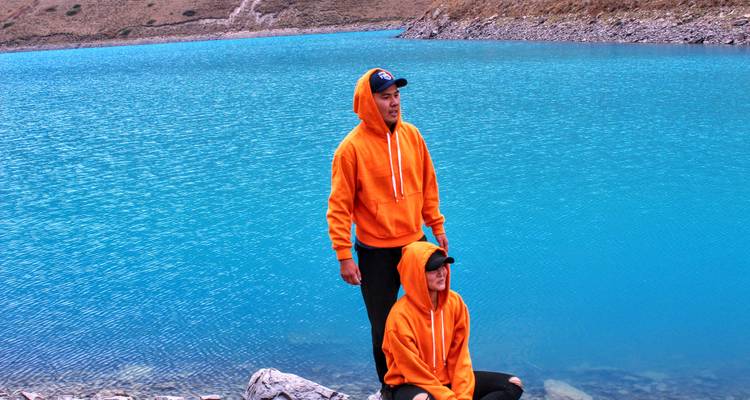Dos excursionistas con sudaderas naranjas a juego posan junto a un lago alpino de un turquesa vívido.