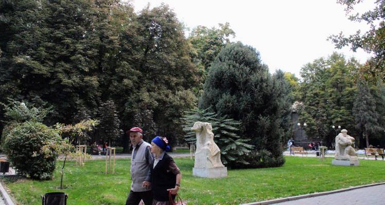 Una pareja de ancianos camina por un parque urbano frondoso con esculturas de piedra y árboles.