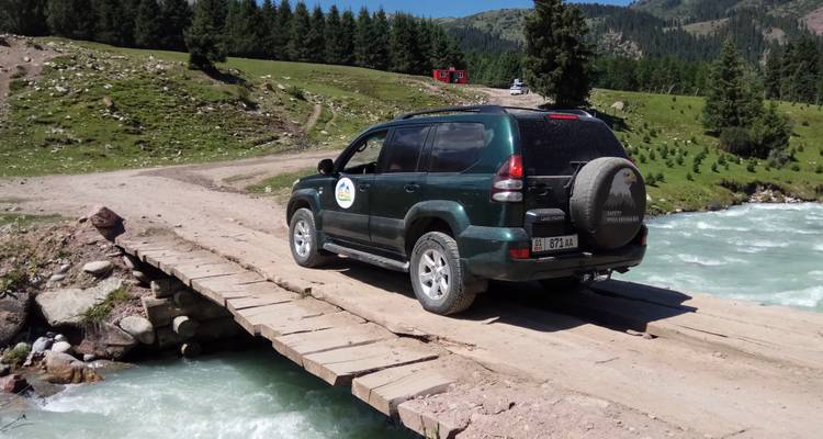 Geländewagen überquert eine Holzbrücke über einen türkisfarbenen Gebirgsfluss.