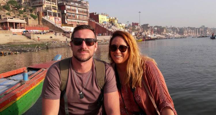 Paar sitzt auf einem Boot mit Varanasi-Ghats und Flussufer-Stadtpanorama im Hintergrund.