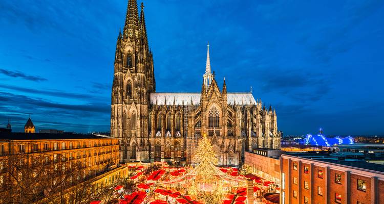 Nächtlicher Blick auf den Kölner Dom, der über einem lebendigen Weihnachtsmarkt mit rotbedachten Ständen erstrahlt.
