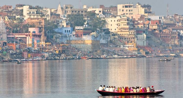Crowded passenger boat on the Ganges with Varanasi ghats and colorful riverside buildings in the background