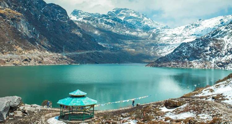 Turquoise Tsomgo Lake surrounded by snow-clad peaks and a small gazebo