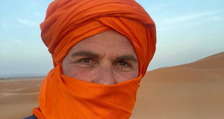 Nahaufnahme eines Reisenden mit leuchtend orangem Turban inmitten sanft geschwungener Sahara-Dünen.