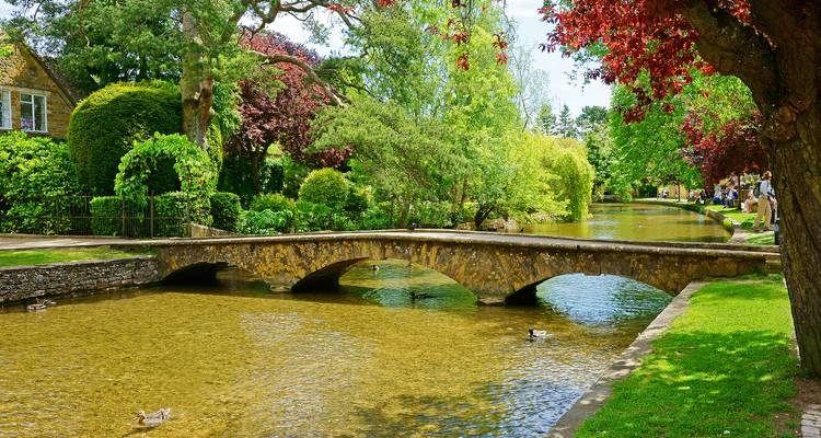 Eine malerische Steinbrücke überquert einen seichten, klaren Bach, der von üppigen Bäumen und dörflichem Grün eingerahmt wird.