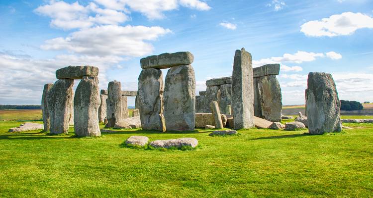Die uralten Steine von Stonehenge stehen auf einer grünen Ebene unter einem strahlend blauen Himmel mit vereinzelten Wolken.