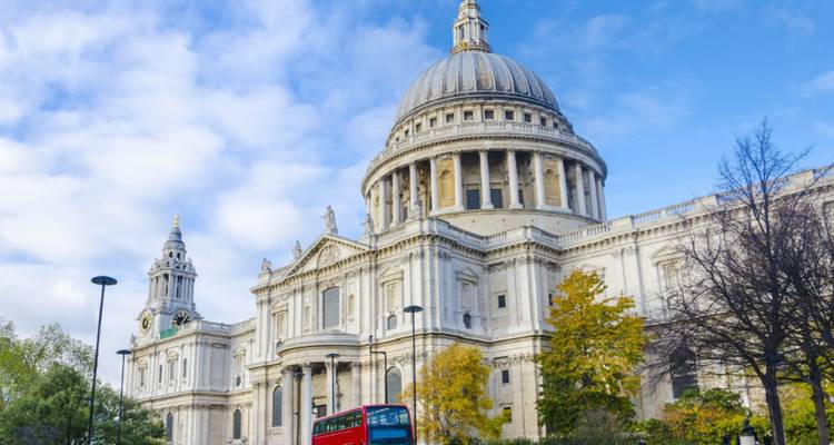 Die St. Paul's Cathedral erhebt sich über herbstliche Bäume, während ein roter Doppeldeckerbus davor vorbeifährt.