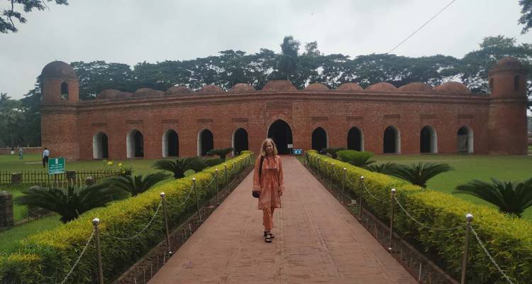 Un visiteur se tient sur un sentier bordé de palmiers menant à la mosquée historique en briques rouges aux soixante dômes