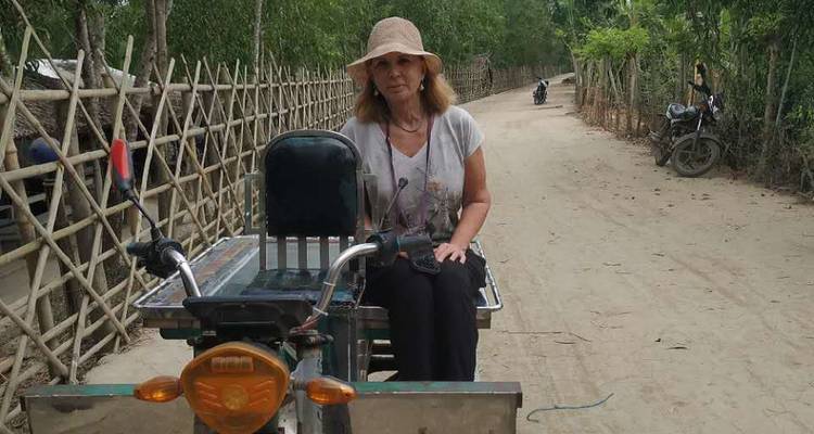 Femme assise sur un petit tuk-tuk à trois roues le long d'une ruelle poussiéreuse de village bordée de clôtures en bambou