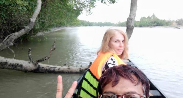 Deux voyageurs dans un petit bateau en bois flottant le long d'une rivière calme de mangrove, l'un faisant un signe de paix vers l'appareil photo.
