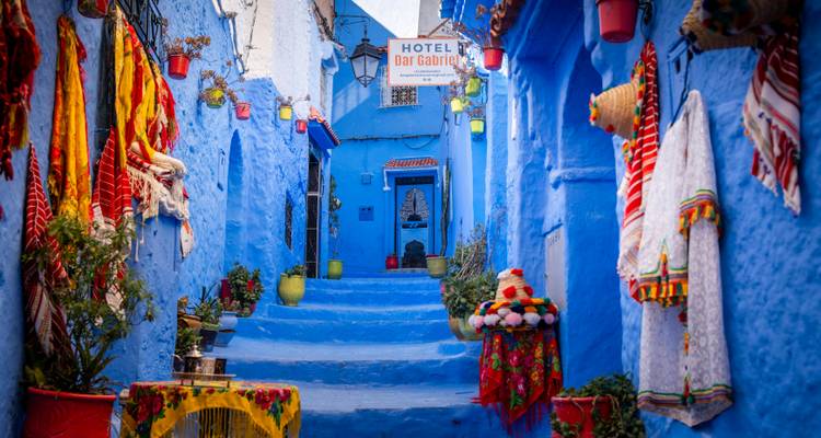 Levendig blauwe steeg in Chefchaouen versierd met kleurrijke textiel, potplanten en een hotelbordje