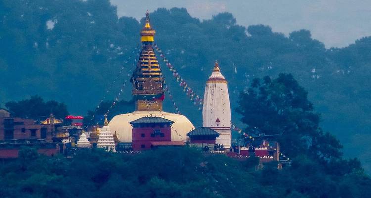 Imagen de teleobjetivo de la estupa Swayambhunath con su aguja dorada y banderas de oración contra una ladera brumosa.