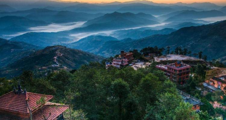 Vista panorámica de cordilleras montañosas escalonadas al amanecer con niebla asentándose en los valles y algunos tejados dispersos en primer plano.