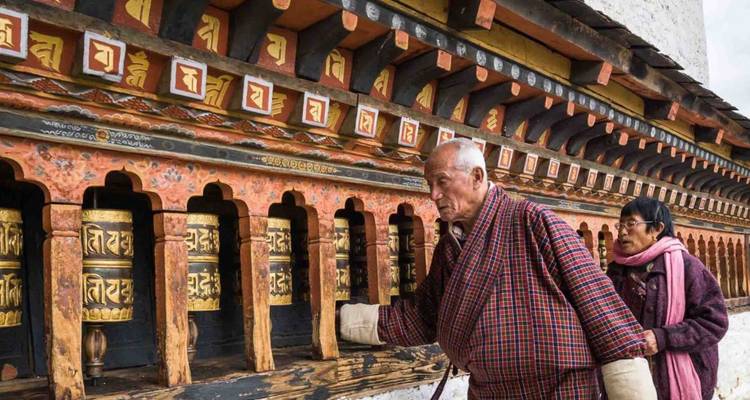 Dos ancianos butaneses vestidos de manera tradicional hacen girar una fila de ruedas de oración ornamentadas empotradas en una pared pintada.
