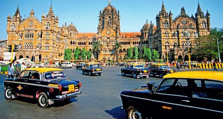 Estación de tren gótica histórica con taxis clásicos negros y amarillos dando vueltas por la plaza concurrida en Mumbai.