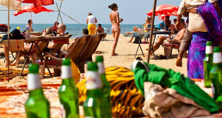 Escena animada de playa goesa con bañistas tomando el sol, sombrillas y botellas de cerveza verdes en primer plano.