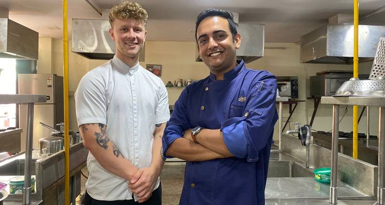 Cocinero sonriente con chaqueta azul está de pie junto a un turista dentro de una cocina industrial de enseñanza.