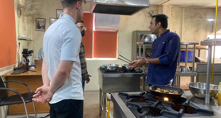 El chef explica técnicas de cocina a dos viajeros dentro de un aula de cocina profesional.
