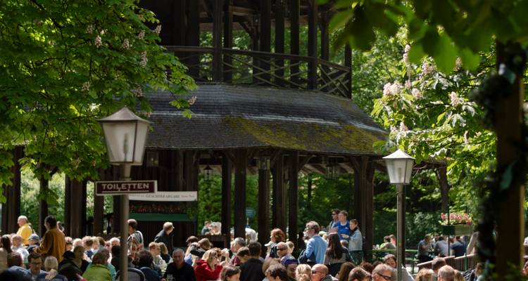 Lebhafte Biergarten-Szene unter dem hölzernen Chinesischen Turm im Münchner Englischen Garten, umgeben von belaubten Bäumen.
