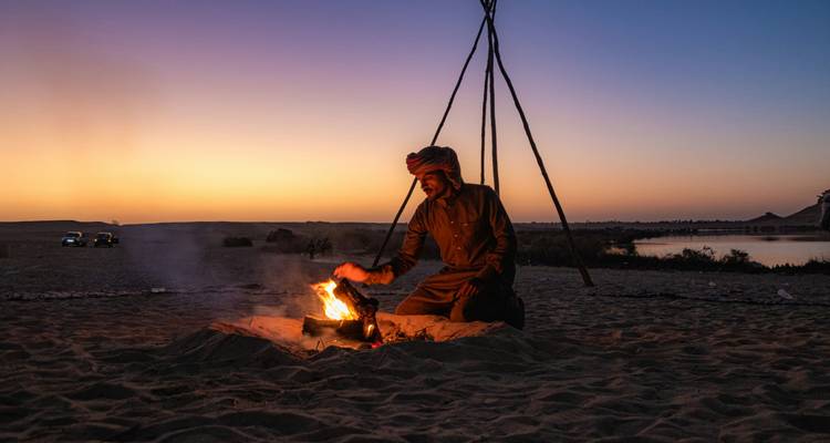 Bedoeïenengids die een klein kampvuur verzorgt op woestijnzand bij schemering met een oranje-paarse zonsonderganglucht.