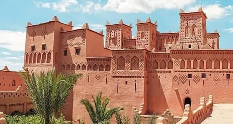 Kasbah élaborée en adobe rose avec des sculptures géométriques ornées et des palmiers se détachant sur un ciel bleu éclatant.