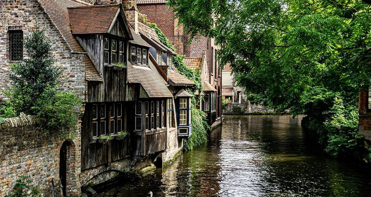 Des maisons médiévales en brique et en bois bordent un canal tranquille encadré par une verdure luxuriante.