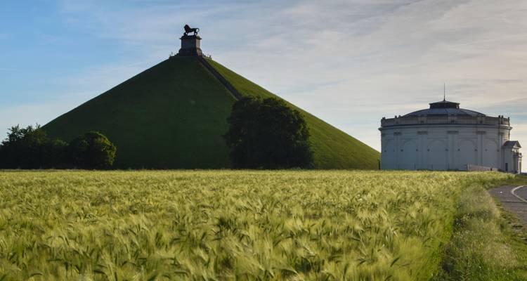 Un monticule conique vert surmonté d'une statue de lion s'élève au-dessus de champs de céréales dorés.
