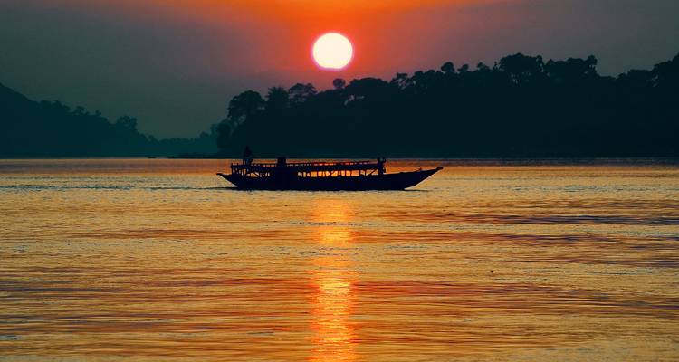 Silhouette eines Holzbootes, das über einen breiten Fluss gleitet, während die Sonne in feurigen Orangetönen untergeht.