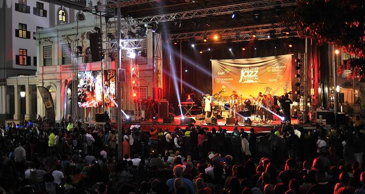 Nächtliches Open-Air-Jazzfestival mit hell erleuchteter Bühne, auftretenden Musikern und einer großen jubelnden Menge.