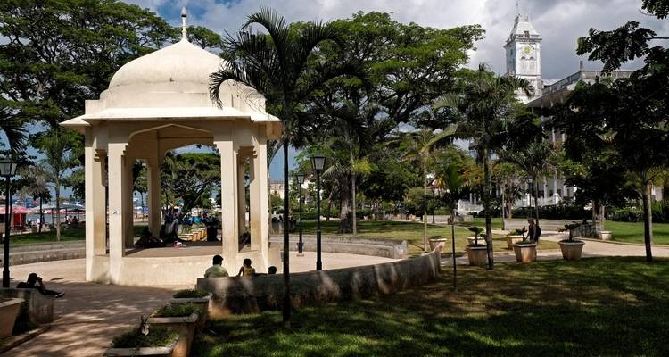 Parque sombreado en Stone Town con un pabellón de cúpula blanca, palmeras altas, visitantes sentados y un edificio histórico con torre de reloj al fondo.