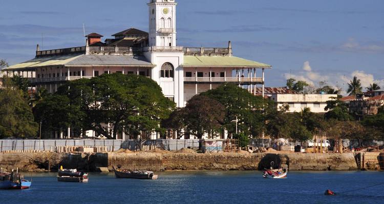 Vista del frente marítimo de un gran edificio colonial con una torre de reloj frente a un puerto azul tranquilo salpicado de pequeños barcos pesqueros.