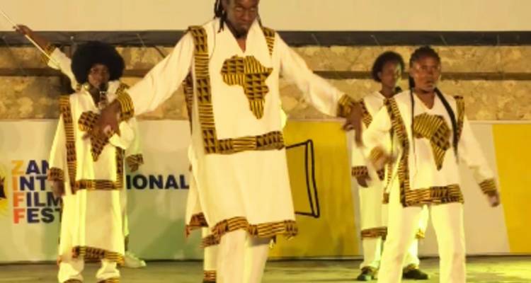 Grupo de bailarines con trajes blancos estampados actuando en un escenario al aire libre bajo focos.