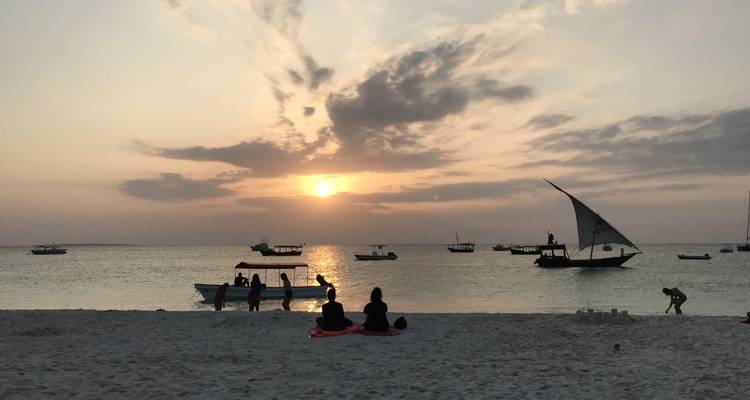 Atardecer dorado sobre un mar en calma con botes tradicionales y bañistas en silueta sobre la arena.