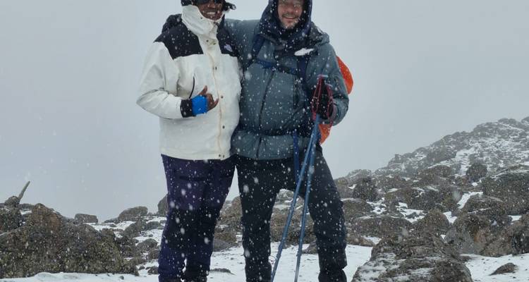 Deux randonneurs en tenue de neige sourient au milieu de flocons qui tombent sur une crête alpine rocheuse.