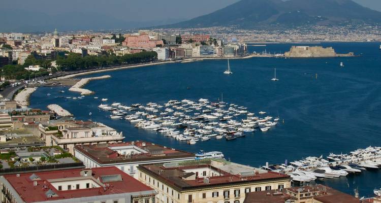 Vue panoramique sur la marina de Naples remplie de yachts blancs, la skyline de la ville et le Mont Vésuve s'élevant en arrière-plan.