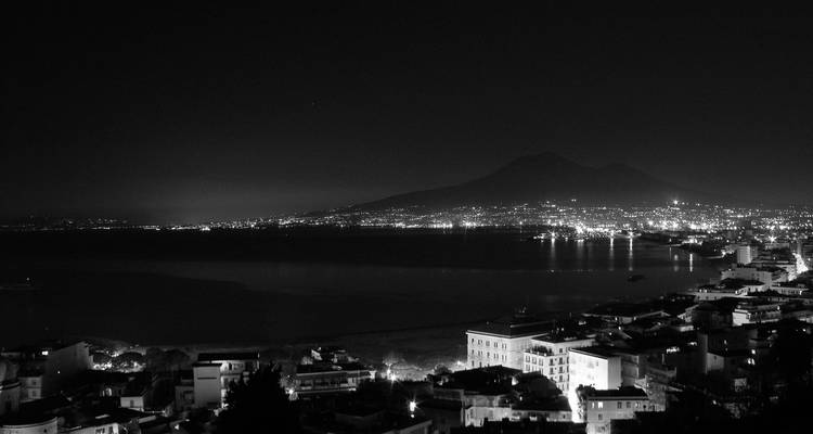 Panorama nocturne en noir et blanc de la baie de Naples avec les lumières de la ville scintillant sous la silhouette du Mont Vésuve.