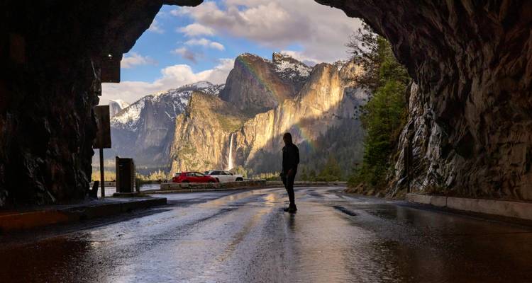Un viajero en silueta se encuentra en un túnel enmarcando el Valle de Yosemite, donde un arcoíris se arquea sobre acantilados de granito.