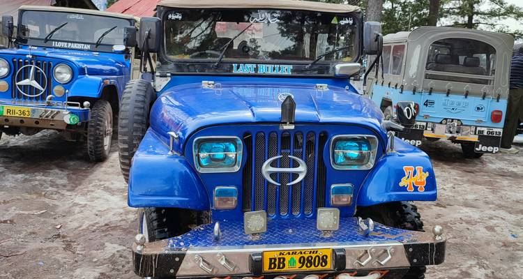 Jeep todoterreno azul brillante estacionado en un terreno de tierra junto a otros jeeps en una región montañosa.