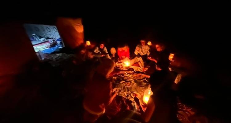 Reizigers zitten op tapijten rond door lantaarns verlichte schotels in een donker woestijnkamp 's nachts.