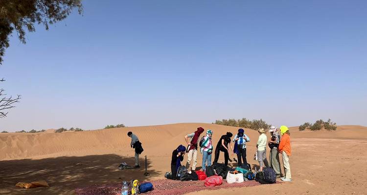 Groep met rugzakken en benodigdheden neemt een pauze naast glooiende zandduinen onder blauwe hemel.