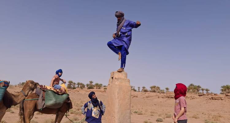 Tuareg-gids in blauwe gewaad balanceert bovenop een steen terwijl toeristen en kamelen toekijken in de woestijn.